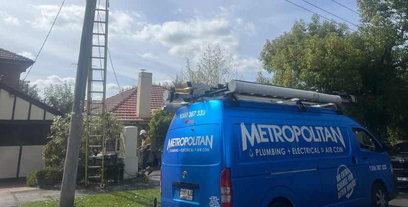 Electrician working outdoors in the front yard of a person's house. At the foreground of the photo is a blue van with Metropolitan Plumbing, Electrical and Air Conditioning branding on it.